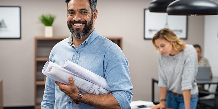 Portrait of multiethnic architect with blueprints in creative office. Mature middle eastern contractor holding roll of architectural projects while looking at camera. Happy latin man in casual standing in meeting room with engineers working in background.